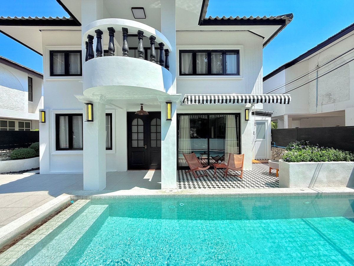 A modern villa exterior features a private swimming pool reflecting sunlight. The structure showcases a covered balcony with decorative railings and large windows. Two lounge chairs are positioned beside the pool, complemented by a patterned tiled surface and lush greenery in the background.