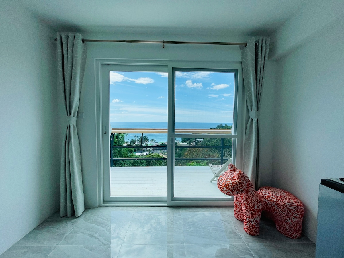 A bright indoor space features large sliding glass doors opening to a balcony with sea views. Soft, neutral curtains frame the entrance, while two vibrant chairs in a coral pattern provide seating. The polished floor reflects light, enhancing the airy feel of the room.
