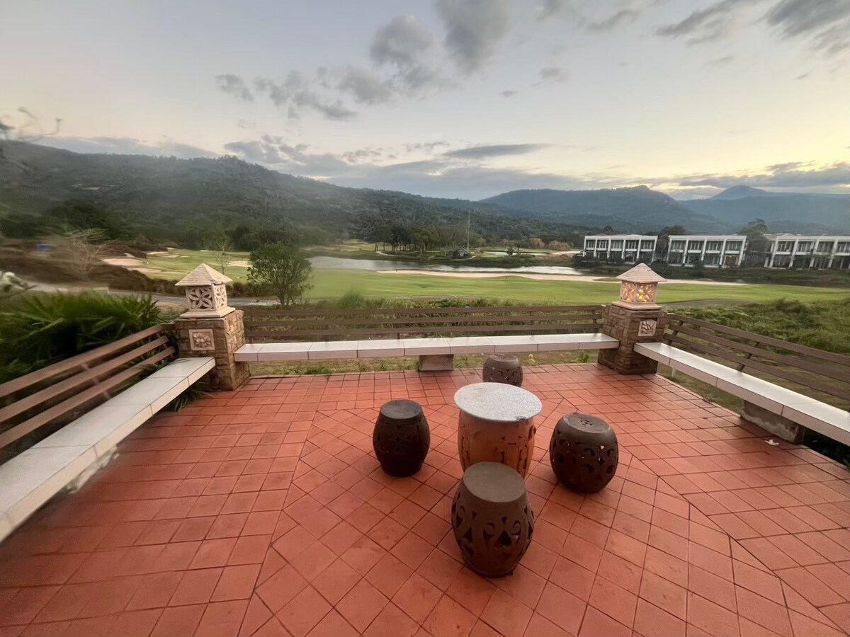 An outdoor seating area features circular stone tables surrounded by low wooden stools. A tiled floor complements the natural landscape view, which includes rolling hills and a golf course in the background. Soft light enhances the serene atmosphere of the space.