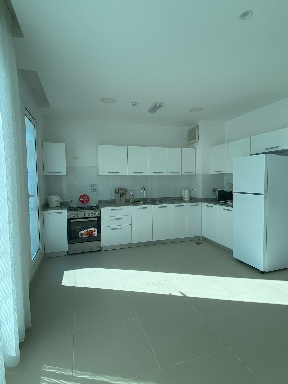 A modern kitchen is presented with sleek white cabinetry and a polished countertop. Essential appliances, including a refrigerator and stove, are included. Natural light floods the space through a large window, creating a bright atmosphere. The tiled floor enhances the contemporary look.