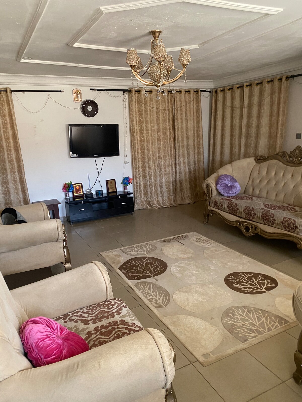 A living room is featured, furnished with ornate seating and a central area rug. A flat-screen LED TV is mounted on the wall, and elegant curtains frame large windows. Decorative items can be seen on a low table, adding to the inviting atmosphere.