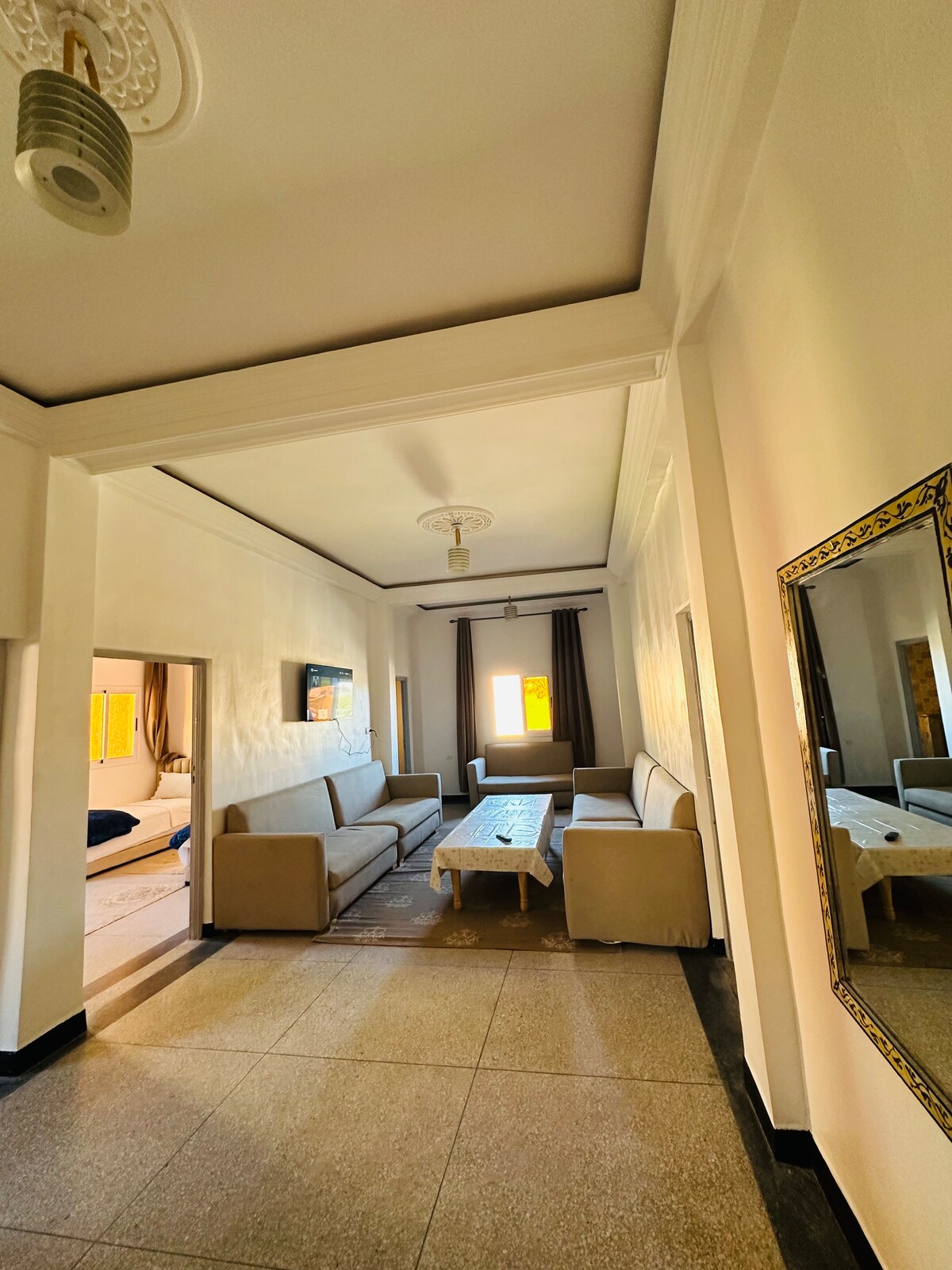 A spacious living area is featured, showcasing a light-colored sofa set arranged around a low coffee table. A wall-mounted television is visible in the background, and natural light enters through a window with curtains. A decorative mirror adds depth to the room.