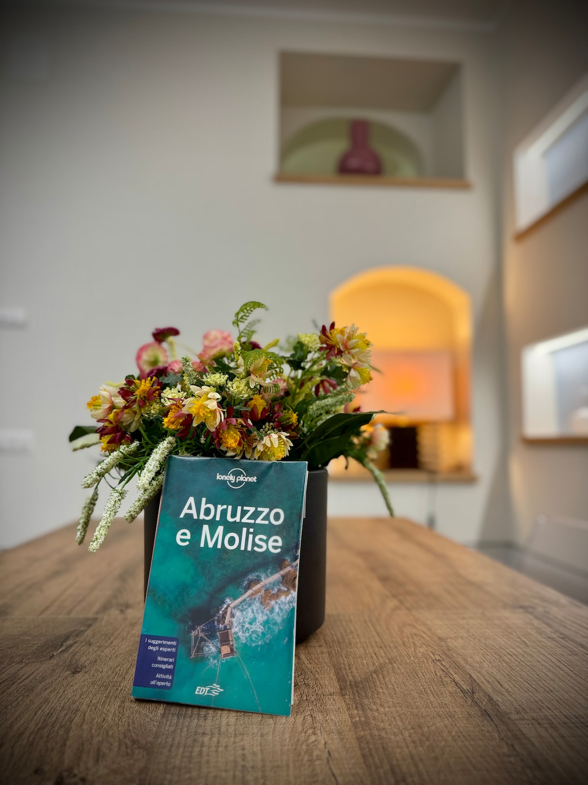 A vibrant arrangement of flowers is positioned in a black vase on a wooden table. A travel guide titled 'Abruzzo e Molise' is displayed prominently, with a softly lit room visible in the background.