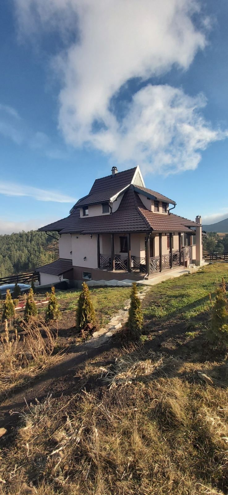 The exterior of a multi-level house is displayed, featuring a combination of wooden and stone elements. Surrounded by green terrain, the property offers balconies and a path leading to the entrance. A scenic backdrop of trees and distant mountains enhances the tranquil setting.
