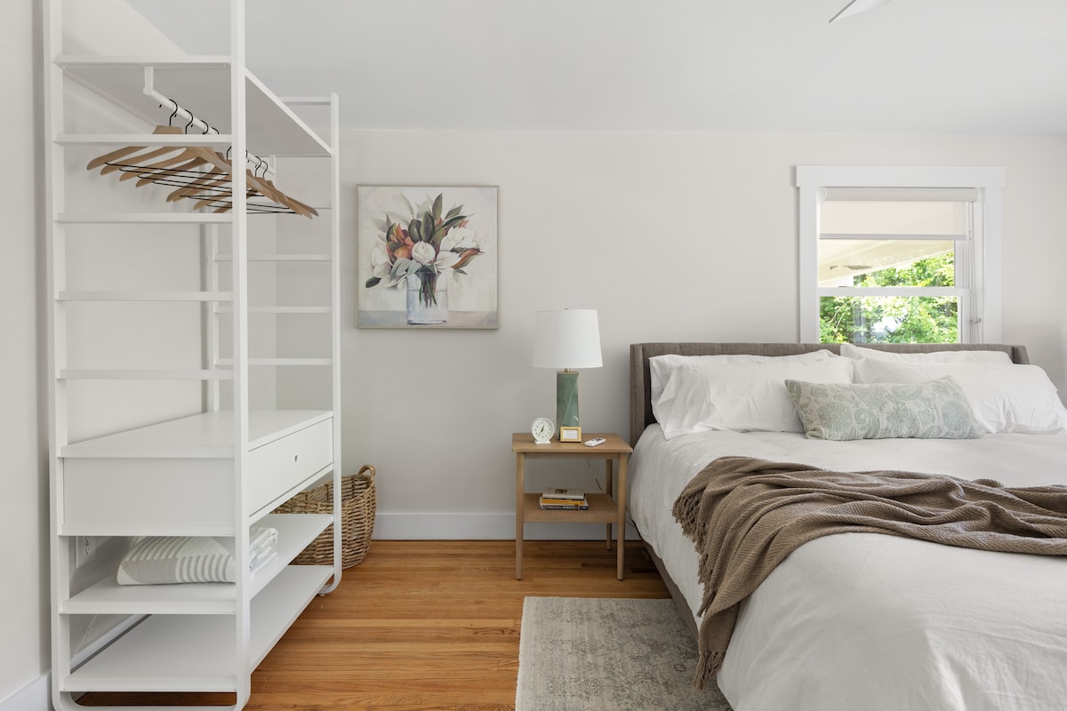 A serene bedroom is highlighted by a spacious bed dressed in soft linens. A minimalist clothing rack and a lamp sit alongside a wooden nightstand. Natural light filters in through a window, illuminating a floral artwork on the wall, creating a calm and inviting space.