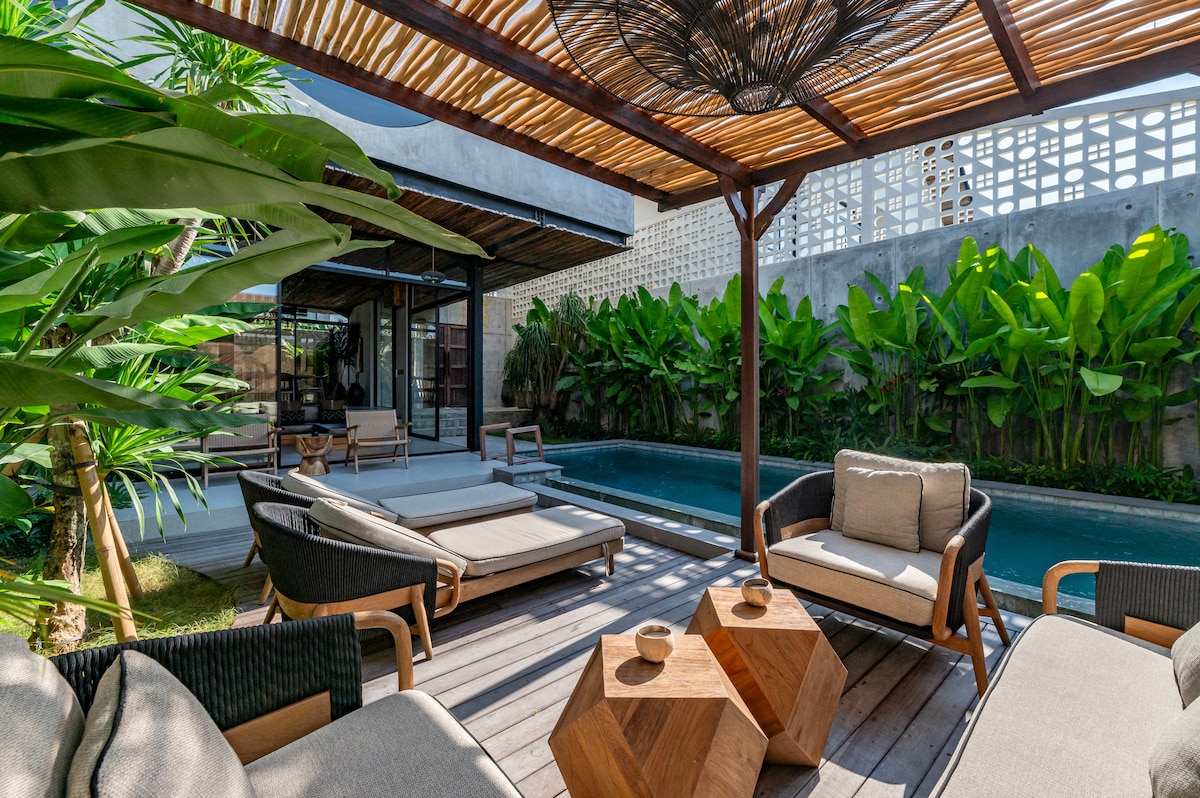 A tranquil outdoor area is presented with sun loungers arranged near a clear swimming pool. A shaded seating arrangement features comfortable cushions on elegant furniture, with a wooden table positioned between two chairs. Lush green plants create a natural border, enhancing the serene setting.