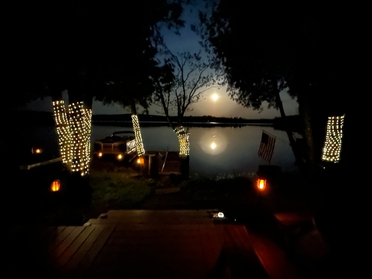 A serene evening view of the lake is captured, highlighted by soft lighting entwined in trees along the water's edge. The reflection of the moon glimmers on the surface, while lanterns provide an ambient glow, enhancing the peaceful atmosphere.