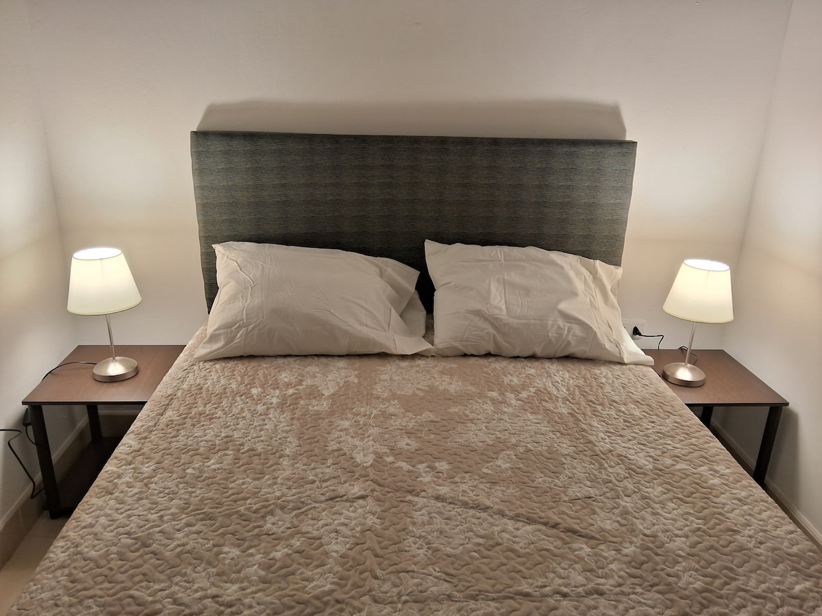 A spacious bed with a soft beige coverlet is centered against a light-colored wall. Two bedside tables are positioned on either side, each holding a lamp with a soft glow, providing gentle illumination to the sleeping area.