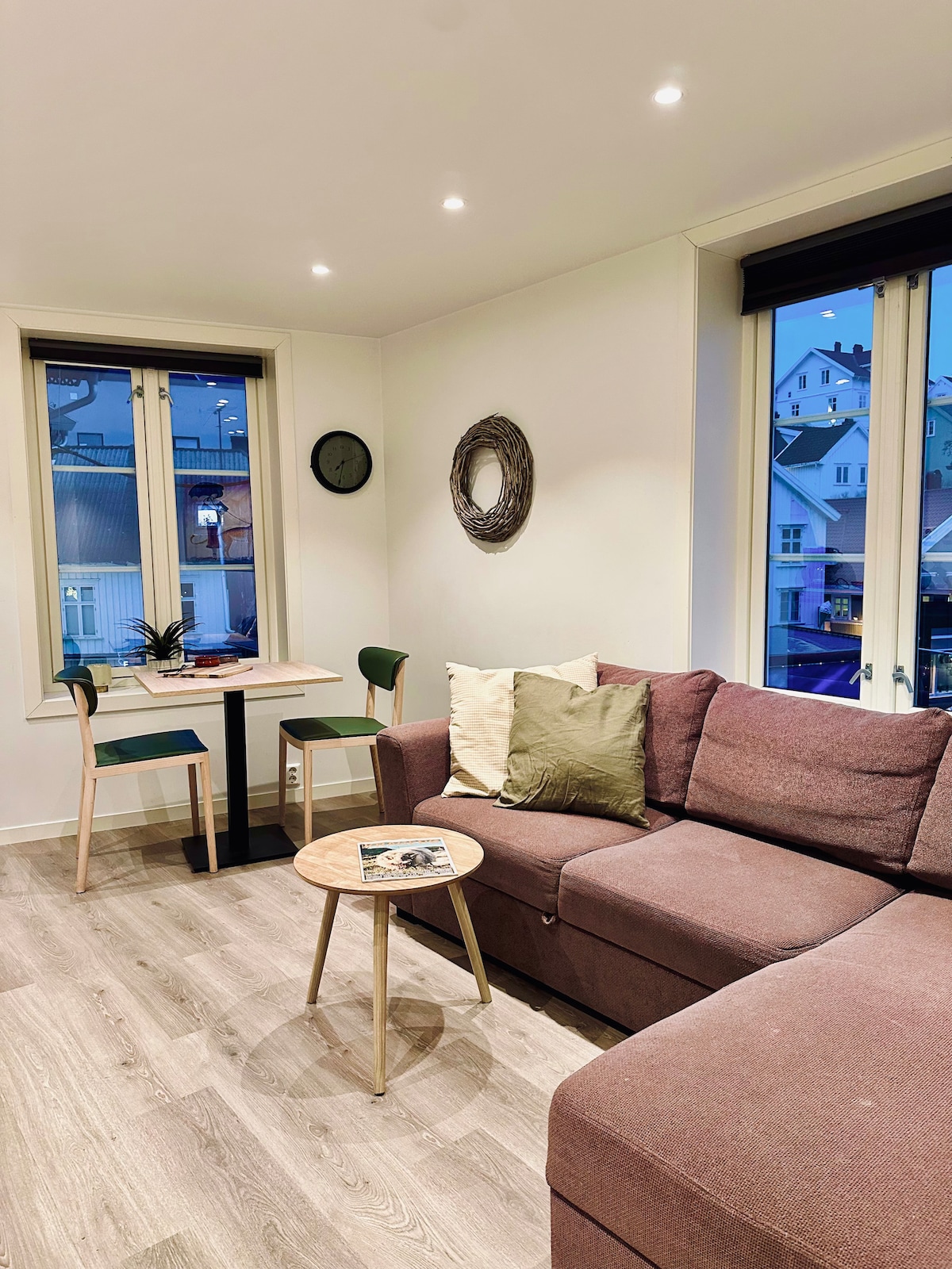 A cozy living area features a modern sofa adorned with neutral pillows, paired with a small round table and two chairs. Natural light enters through large windows, illuminating the space with a soft glow, while a decorative wall mirror adds depth.