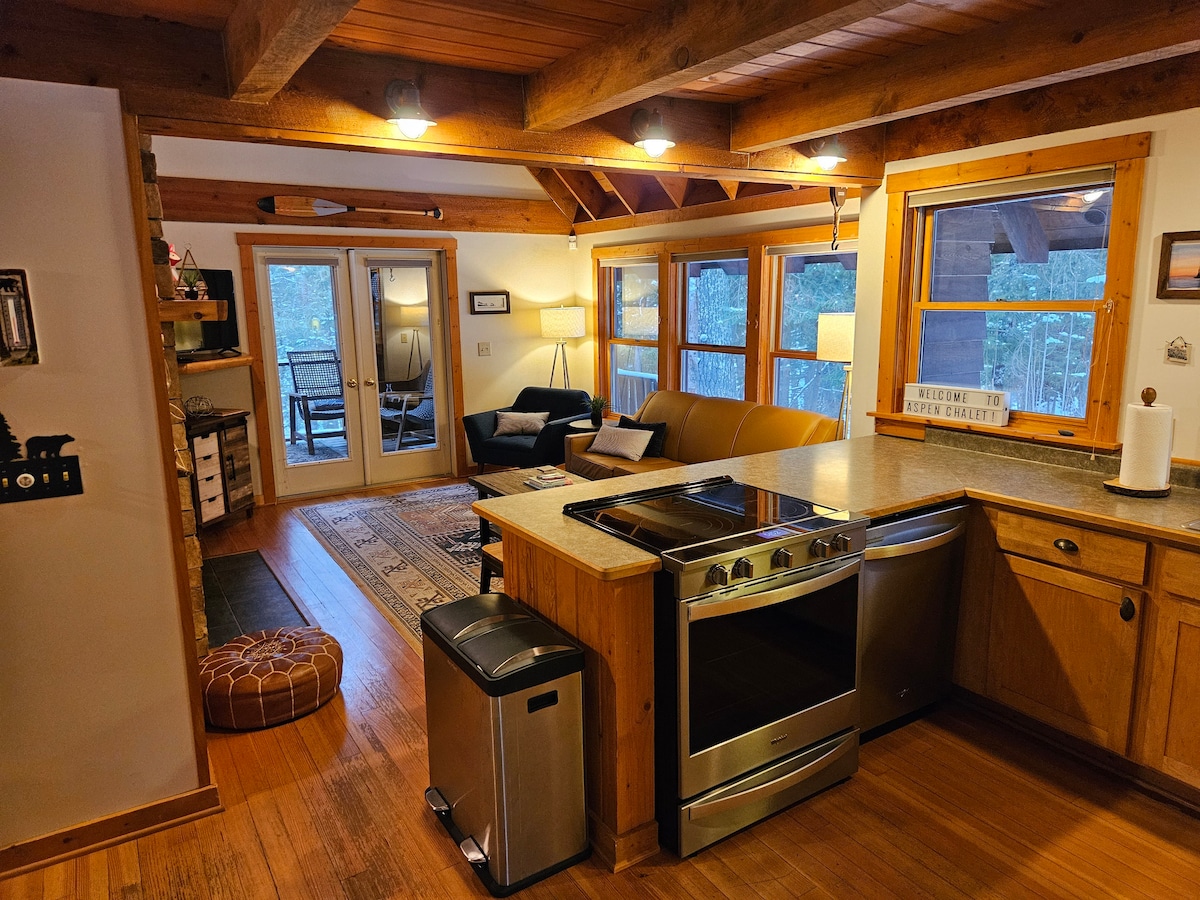 An open kitchen area features wooden cabinetry and stainless steel appliances. A stone countertop provides a workspace facing the living area. Natural light filters through large windows, illuminating a cozy seating space with a couch and a tasteful area rug. Sliding glass doors lead to the outside.
