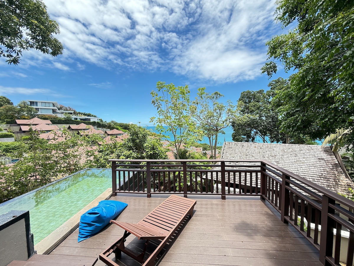 A spacious terrace with wooden flooring features a sun lounger positioned beside a private pool. The pool reflects the vibrant greenery surrounding the area. A scenic view of the sea and distant buildings unfolds against a sky filled with scattered clouds.