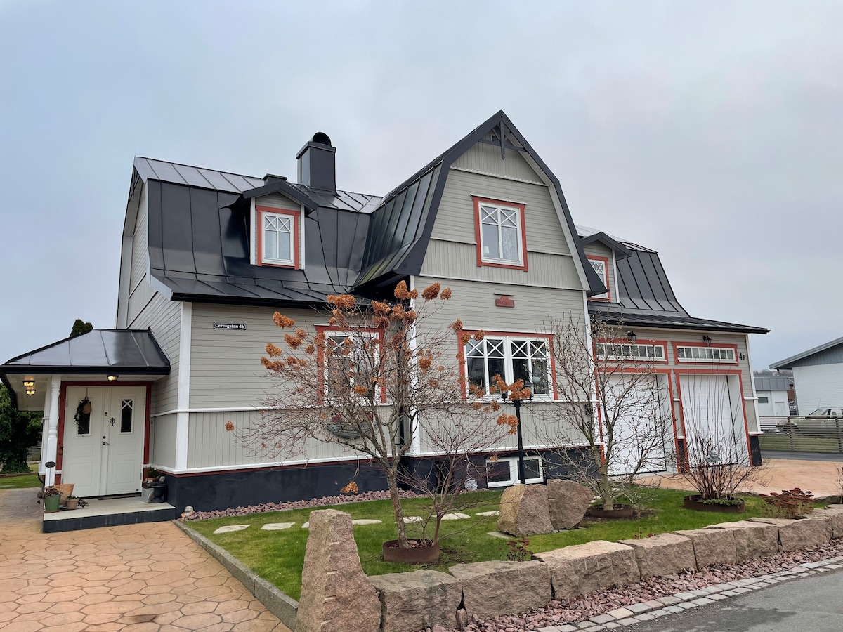 A charming older house is displayed with a unique roof design, featuring a combination of dark and light exterior colors. The front yard includes neatly arranged stones and a small tree, while a driveway leads to the garage on one side.