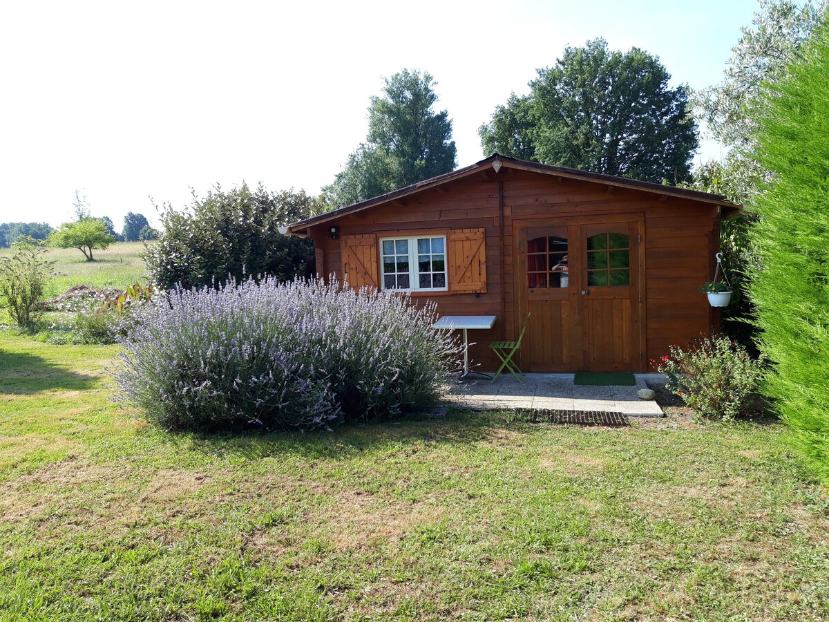 A charming wooden cabin is surrounded by lush greenery and blooming lavender. The cabin features large glass windows with wooden shutters. A small outdoor seating area with a green table and chair is positioned on a stone pathway leading to the entrance.