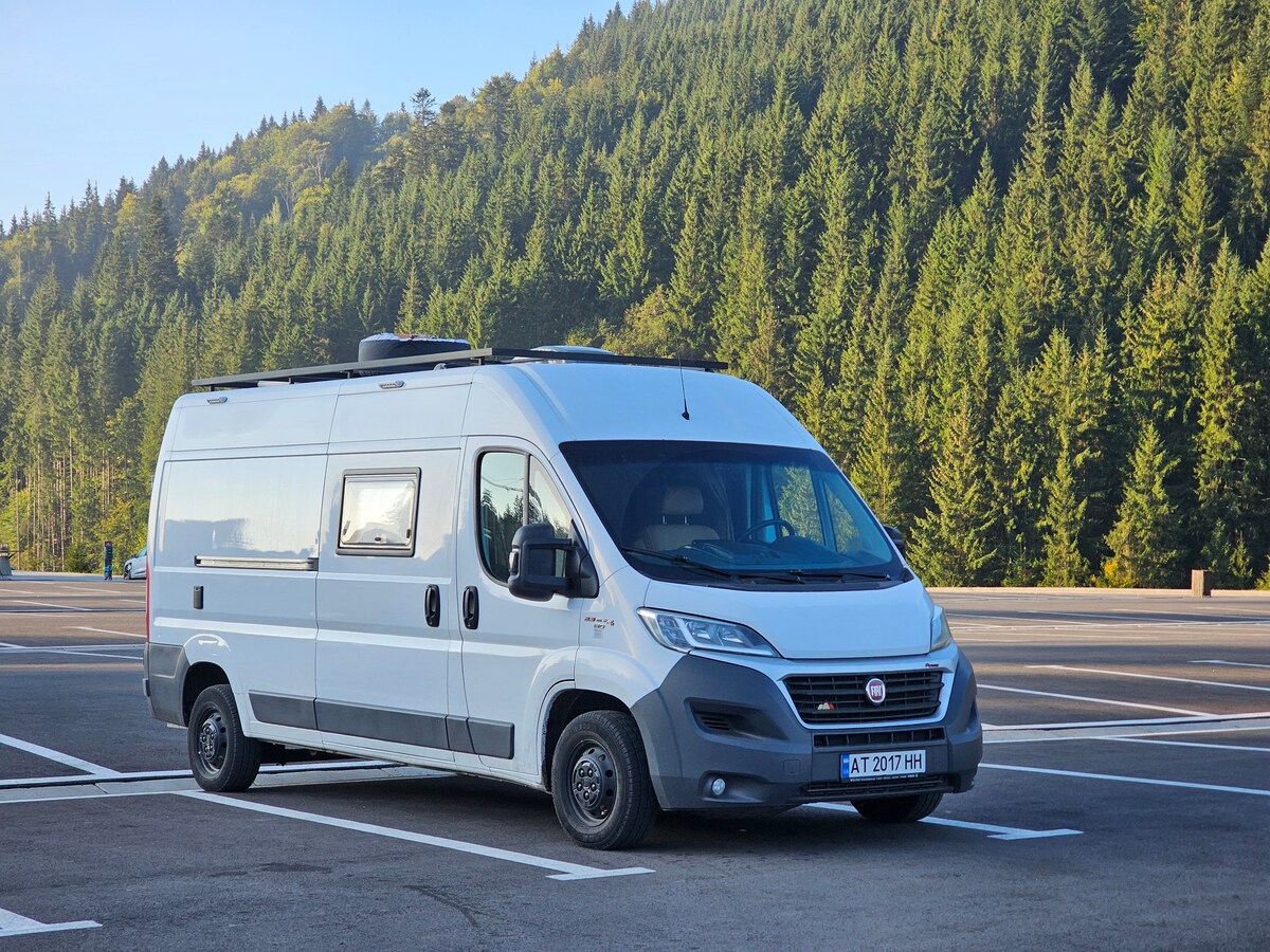 A silver van is parked in a spacious lot, surrounded by tall green trees. The vehicle features large windows and a streamlined exterior, highlighting its design meant for travel. The background includes a serene hillside, providing a sense of peace and adventure.