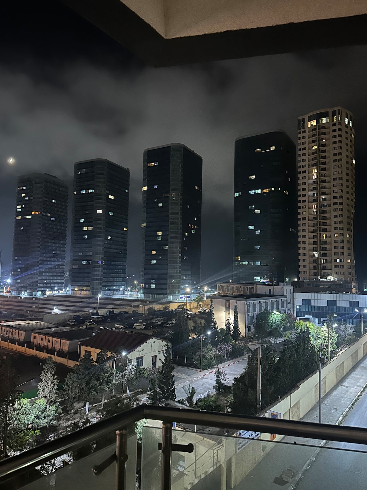 A night view from a balcony highlights a skyline dominated by tall modern buildings, with a mix of glass and concrete structures. Soft clouds drift through the sky, and city lights create a vibrant atmosphere below. Trees and streets are faintly visible in the foreground.