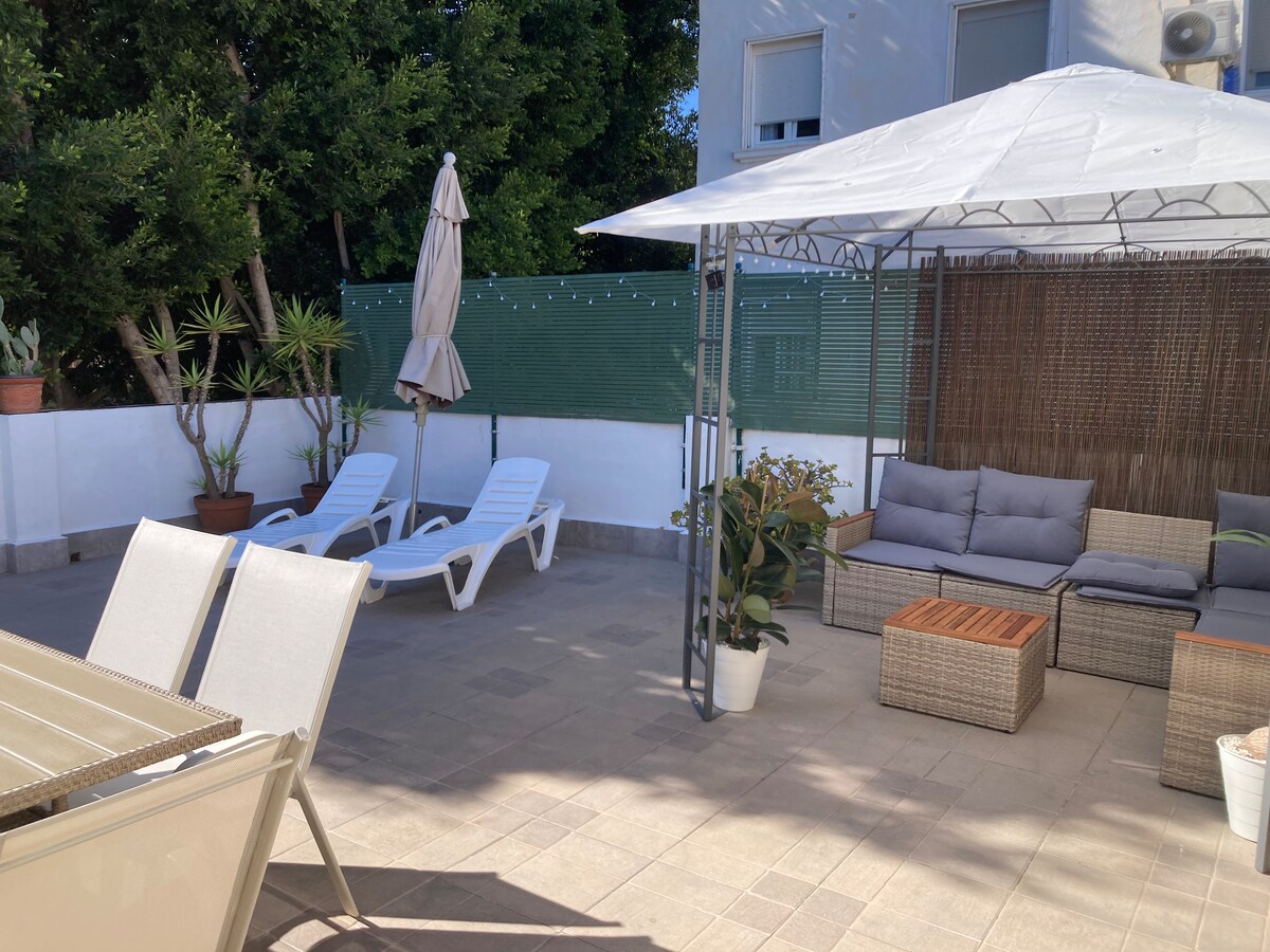 A private terrace area is shown, featuring comfortable seating beneath a canopy. Two loungers are positioned for sunbathing, surrounded by green plants. A dining table with chairs is visible, creating a space for outdoor gatherings and relaxation.