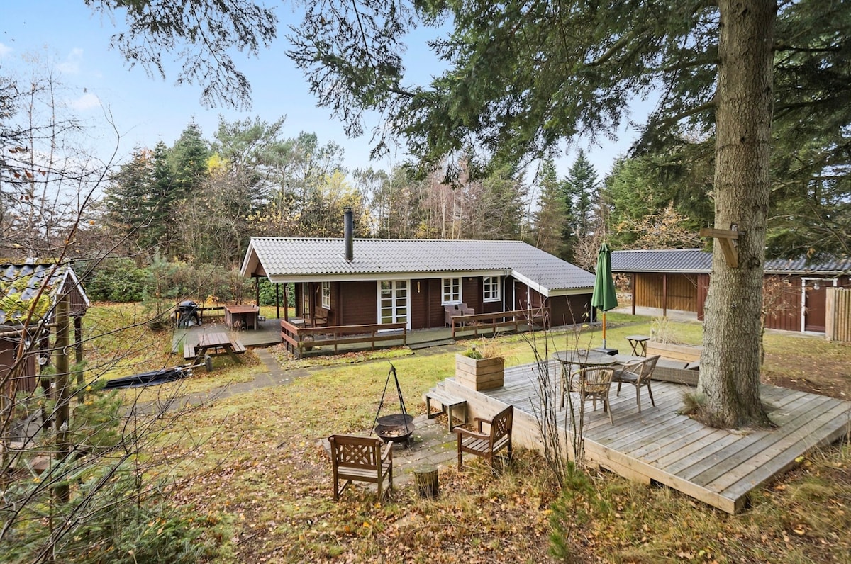 The outdoor area features a spacious wooden deck surrounded by trees. A fire pit is situated nearby, accompanied by several seating arrangements. Additionally, a covered terrace extends from the main building, providing ample space for relaxation in a natural setting.