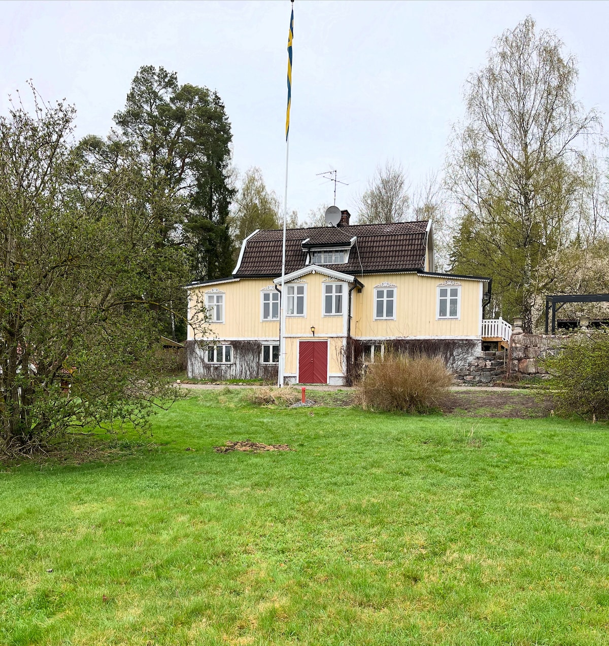 A two-story yellow house with white trim is featured, surrounded by green grass and trees. A red door is visible at the front entrance, with windows on both levels providing natural light. The property includes a flagpole, enhancing the outdoor setting.