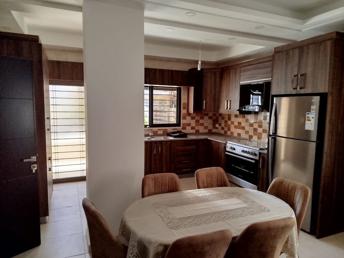 A modern kitchen area features dark wooden cabinets and a tiled backsplash in warm tones. An organized dining table with upholstered chairs is positioned centrally, while a large window allows natural light to illuminate the space.
