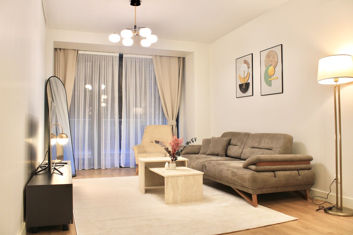 A spacious living area showcases a comfortable sofa and armchair facing a minimalist coffee table. Tall windows are dressed in sheer curtains, allowing natural light to fill the room. Nearby, a lamp and decorative artwork enhance the relaxing ambiance.