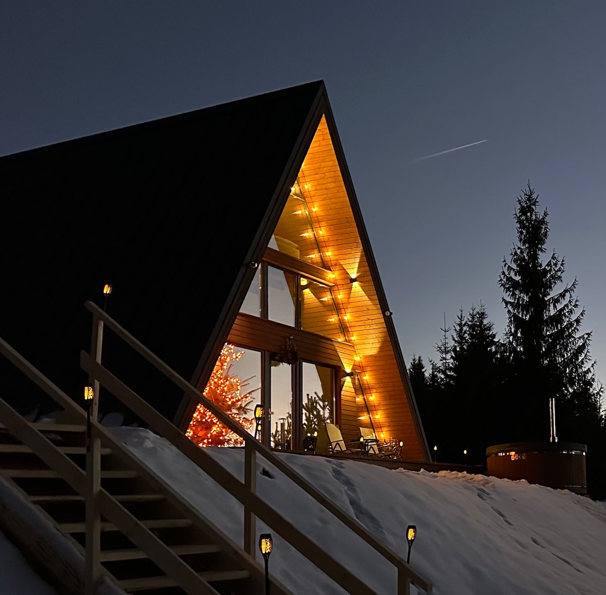 An A-frame cabin stands against a twilight sky, illuminated by warm lights. Snow covers the ground, and a decorated tree is positioned near the entrance. Steps lead to the cabin, while tall trees surround the property, contributing to a serene atmosphere.