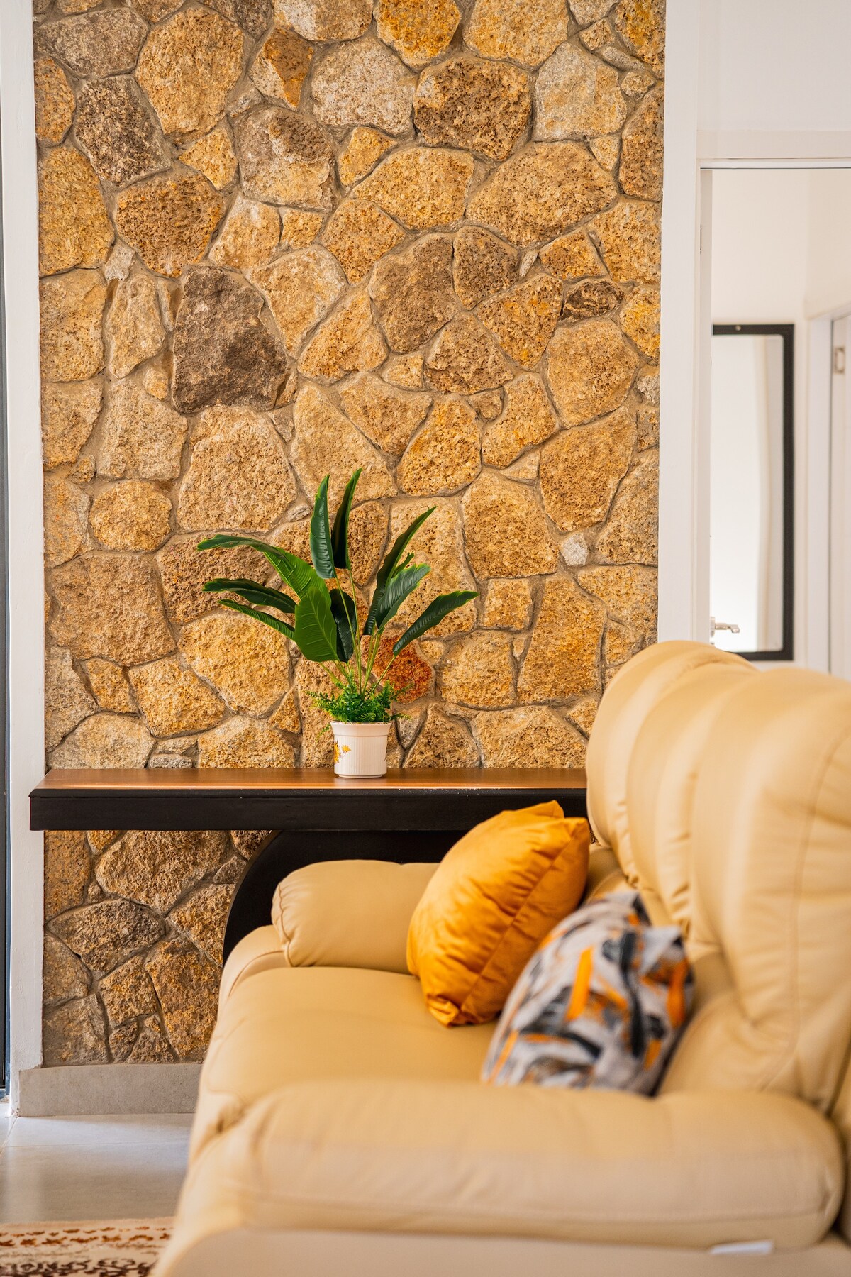 A neutral-toned sofa with decorative cushions is positioned against a textured stone wall. A small potted plant adds a touch of greenery on a dark wooden table nearby. The space is brightened by ample natural light from the adjoining room.