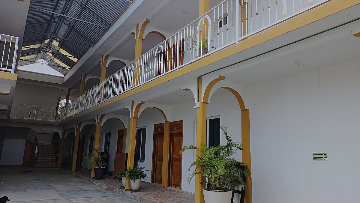A pleasant walkway is lined with several doors leading to individual rooms. A decorative balcony adds dimension to the upper level, accompanied by simple planters. The open area is shaded, with a skylight allowing natural light to illuminate the space.