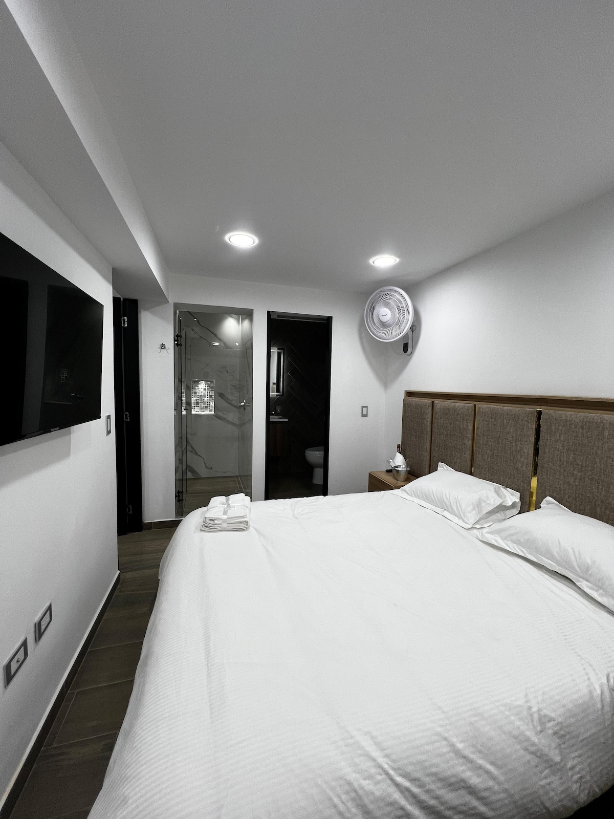 A well-appointed bedroom features a spacious bed adorned with crisp white linens. A wall-mounted television is positioned across from the bed. A glass-enclosed bathroom is accessible in the background, showcasing modern design elements.