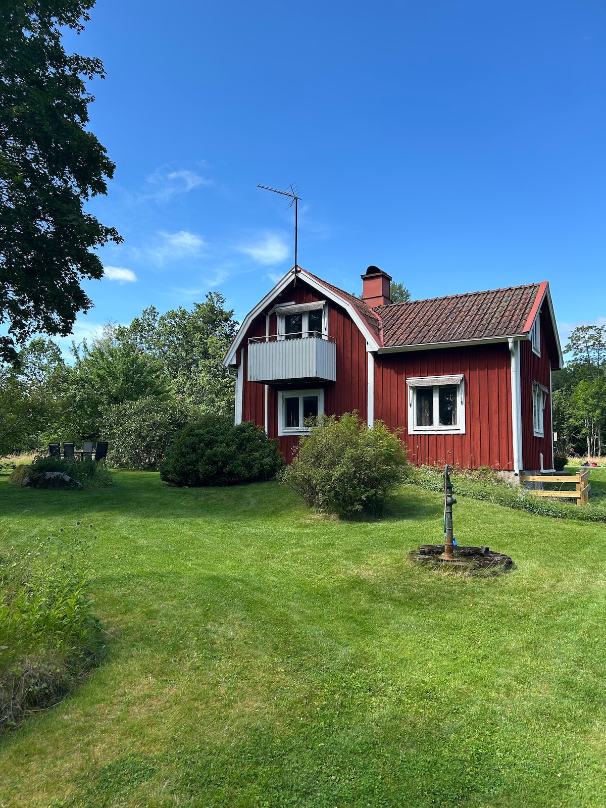 A charming red house is nestled among greenery, featuring a sloping roof and a prominent balcony. The lawn is vibrant, with various shrubs and trees providing a natural backdrop. A well is visible in the garden, adding to the rustic appeal of the location.