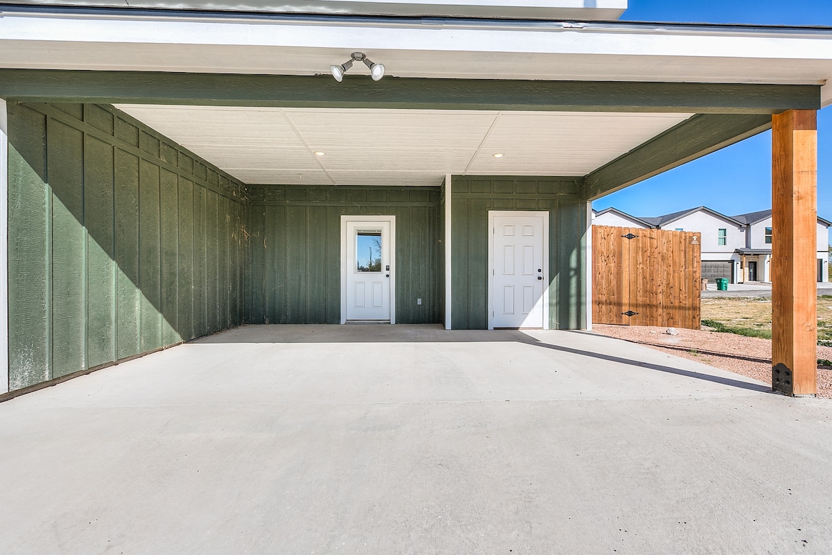 A covered outdoor space is shown, featuring a concrete surface and green siding. Two doors are visible on one wall, leading to different areas. Natural light illuminates the area, highlighting the clean, minimalist design. A wooden fence can be seen in the background.