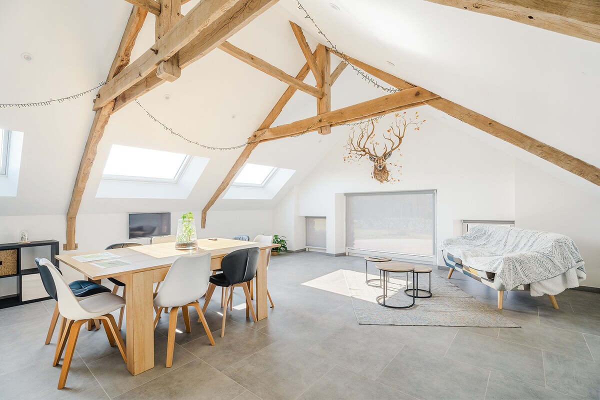 A spacious living area features high ceilings adorned with exposed wooden beams. A large dining table is set with chairs, while a comfortable sleeping area is shown with a neatly made bed and natural light streaming through Velux windows. A cozy seating arrangement is visible, enhancing comfort.