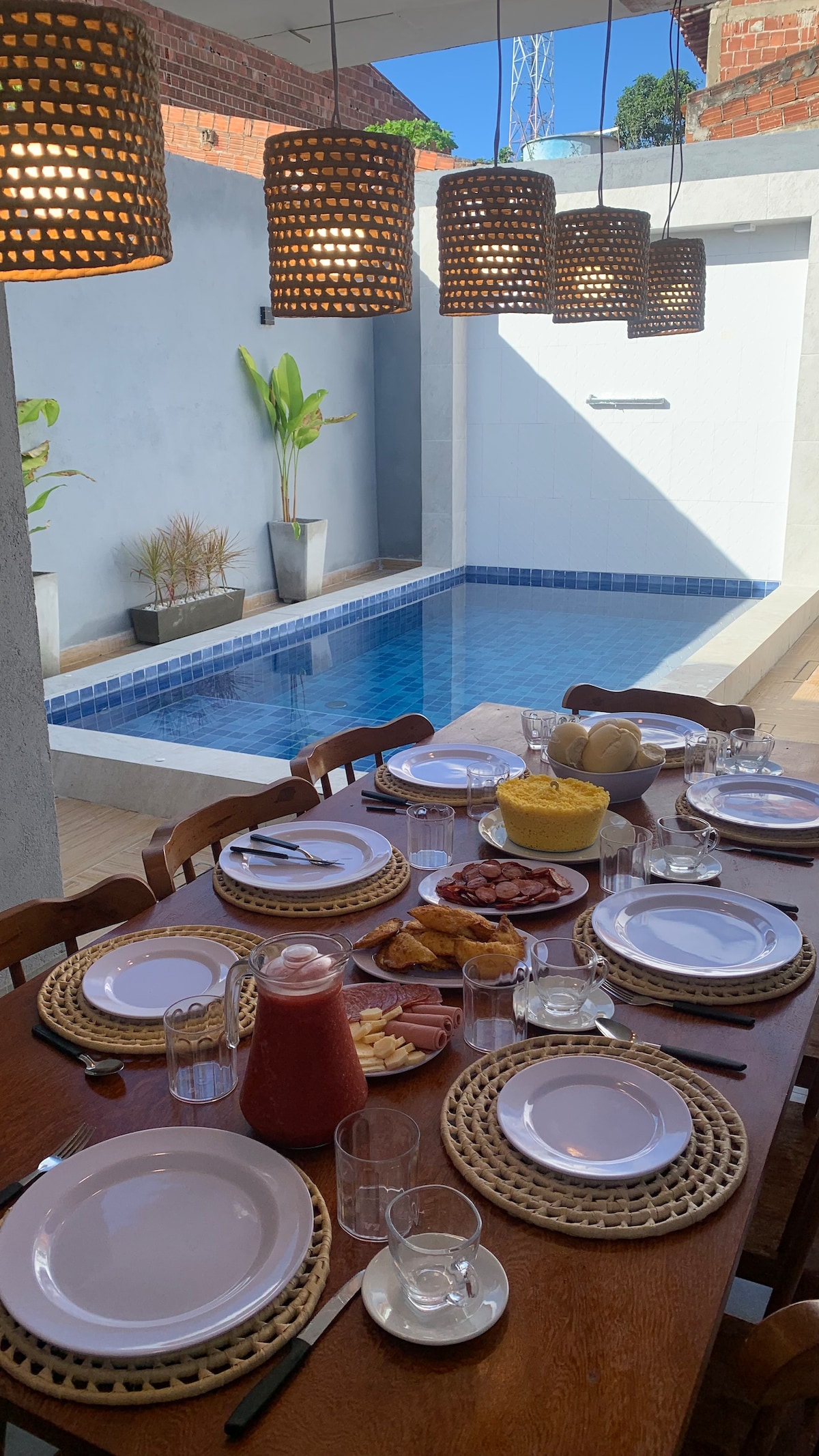 A dining table is set with white plates, glassware, and woven placemats, situated near a gleaming swimming pool. Overhead, pendant lights cast a warm glow. Potted plants enhance the outdoor ambiance, offering a serene space for meals.