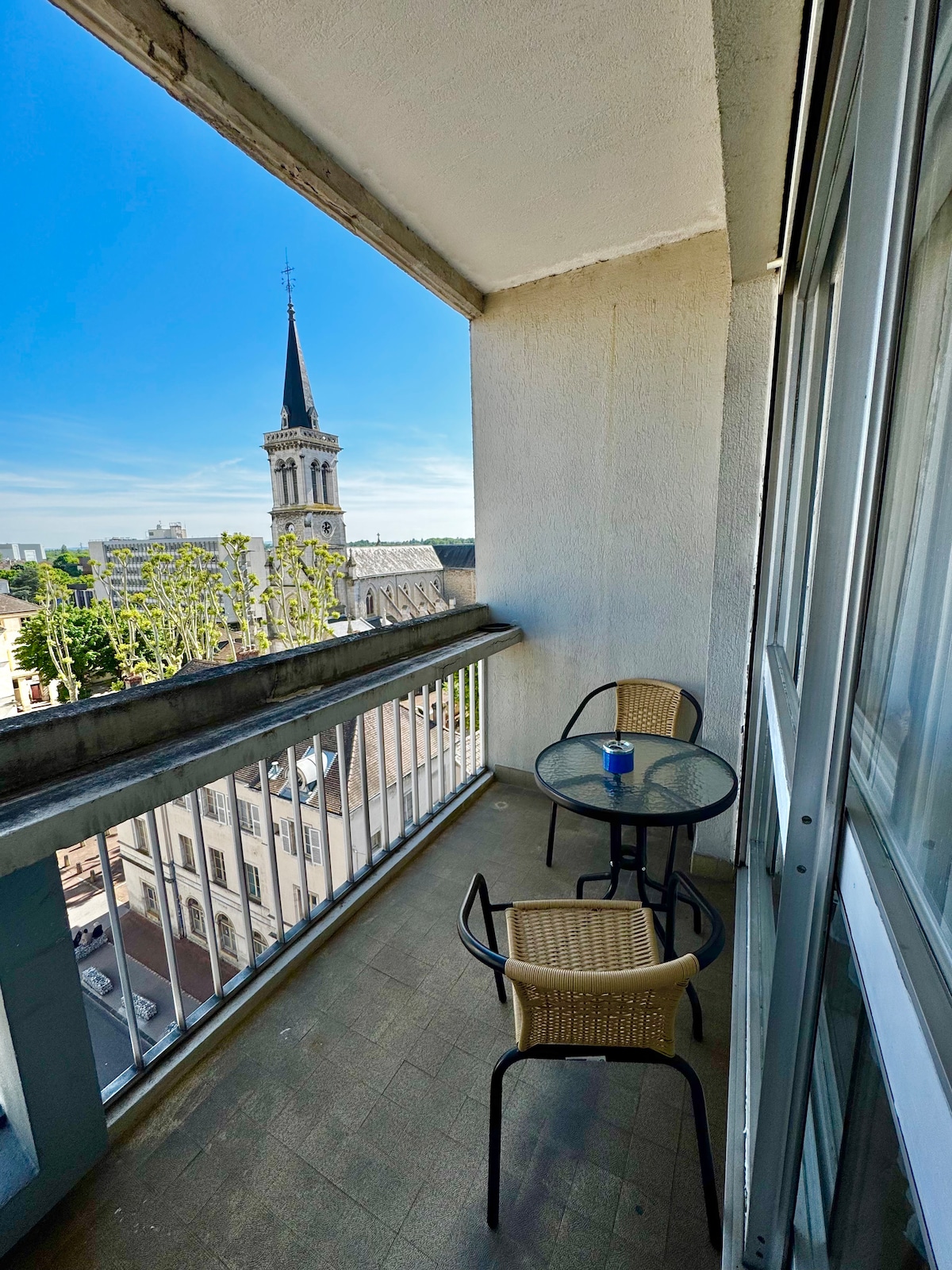A balcony is featured, overlooking a city view that includes a church steeple. Two chairs and a glass table are present, providing a space for relaxation. The area is brightened by natural light, with blue sky visible beyond the railing.