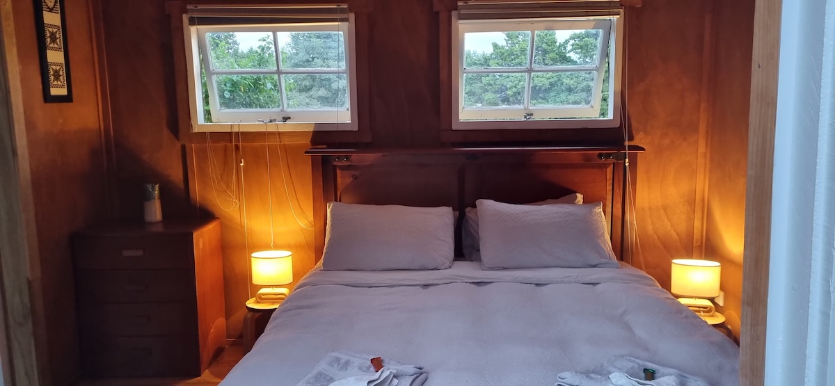 A cozy sleeping area features a comfortable bed with light bedding and two bedside lamps providing a warm glow. Windows on either side allow natural light to enhance the wooden interior and offer views of the greenery outside.