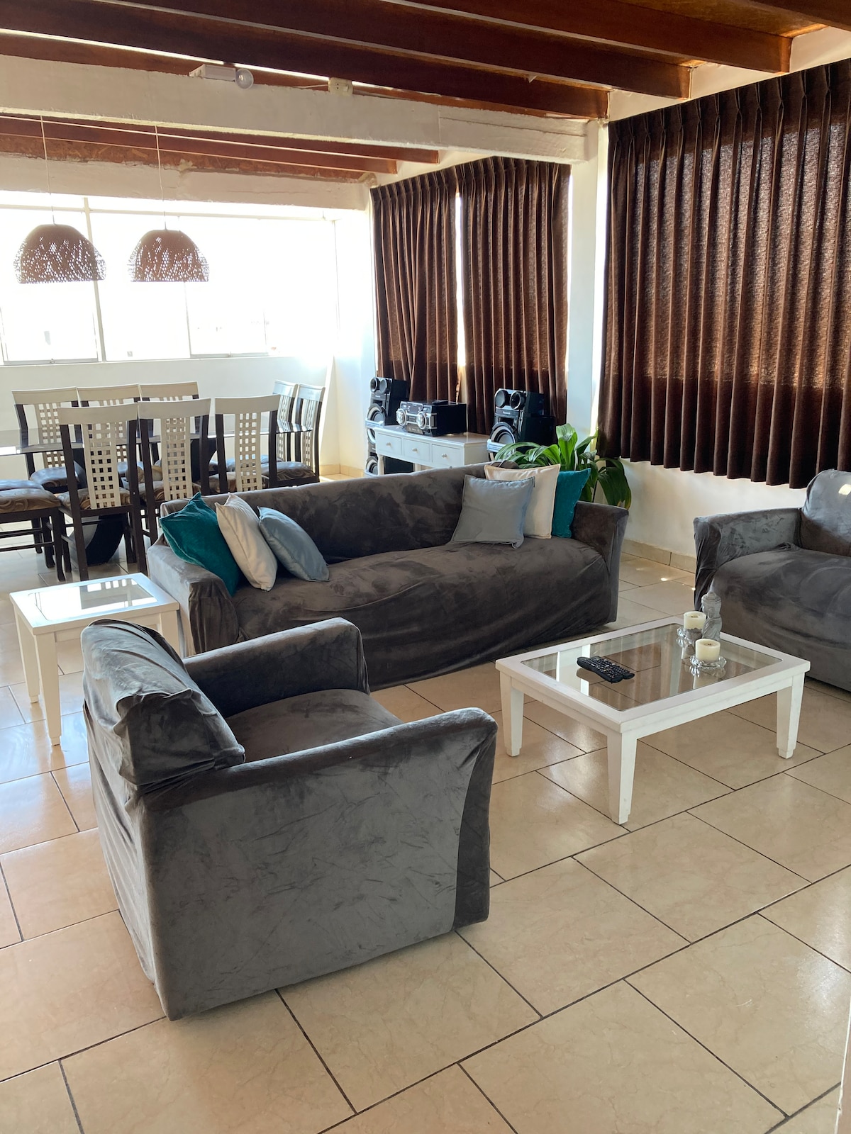 A spacious seating area features plush gray sofas arranged around a white coffee table. Natural light floods the room through large windows, draped with dark curtains. In the background, a dining table with chairs is visible, along with decorative elements that enhance the space.