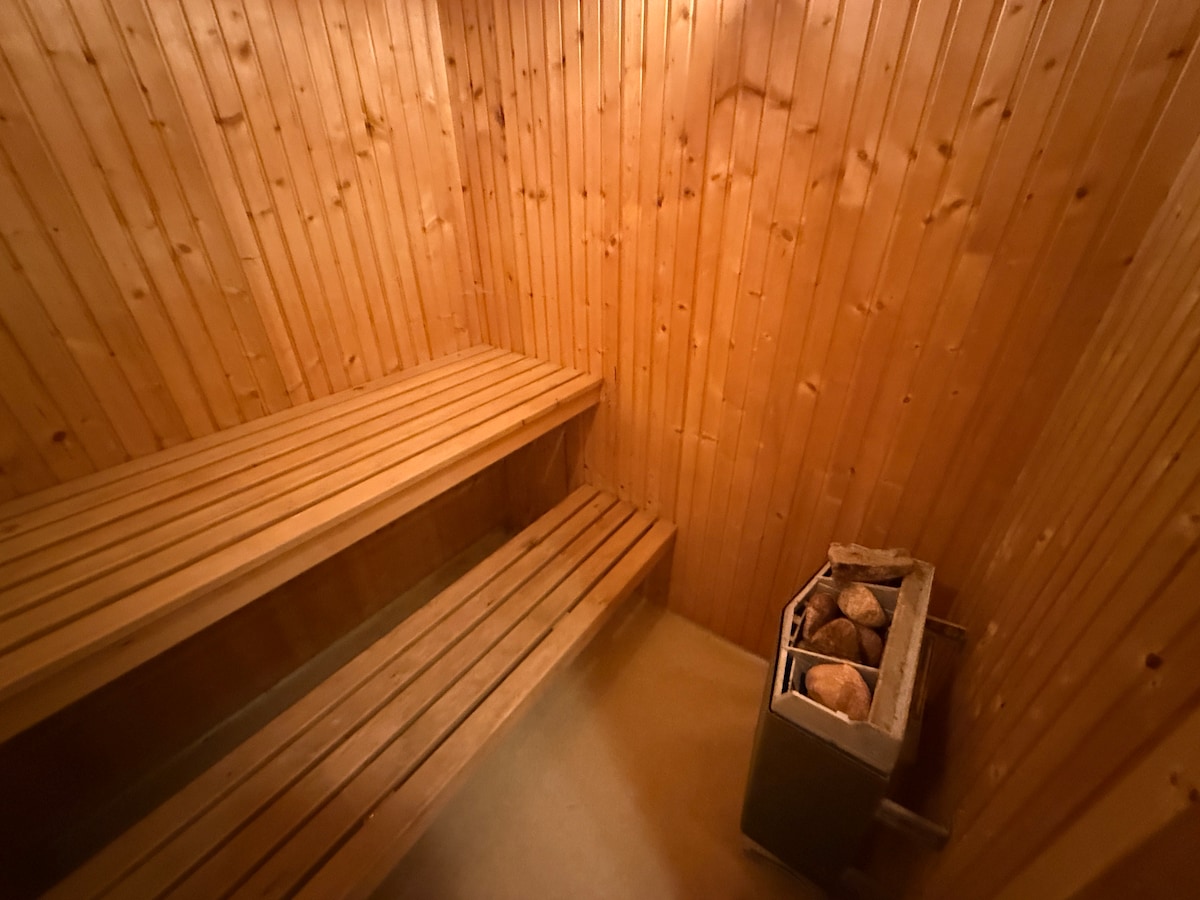 The sauna features warm wooden paneling with built-in benches. A stone heater, positioned against the wall, is visible, providing a cozy ambiance. The warm tones of the wood create an inviting and relaxing environment.