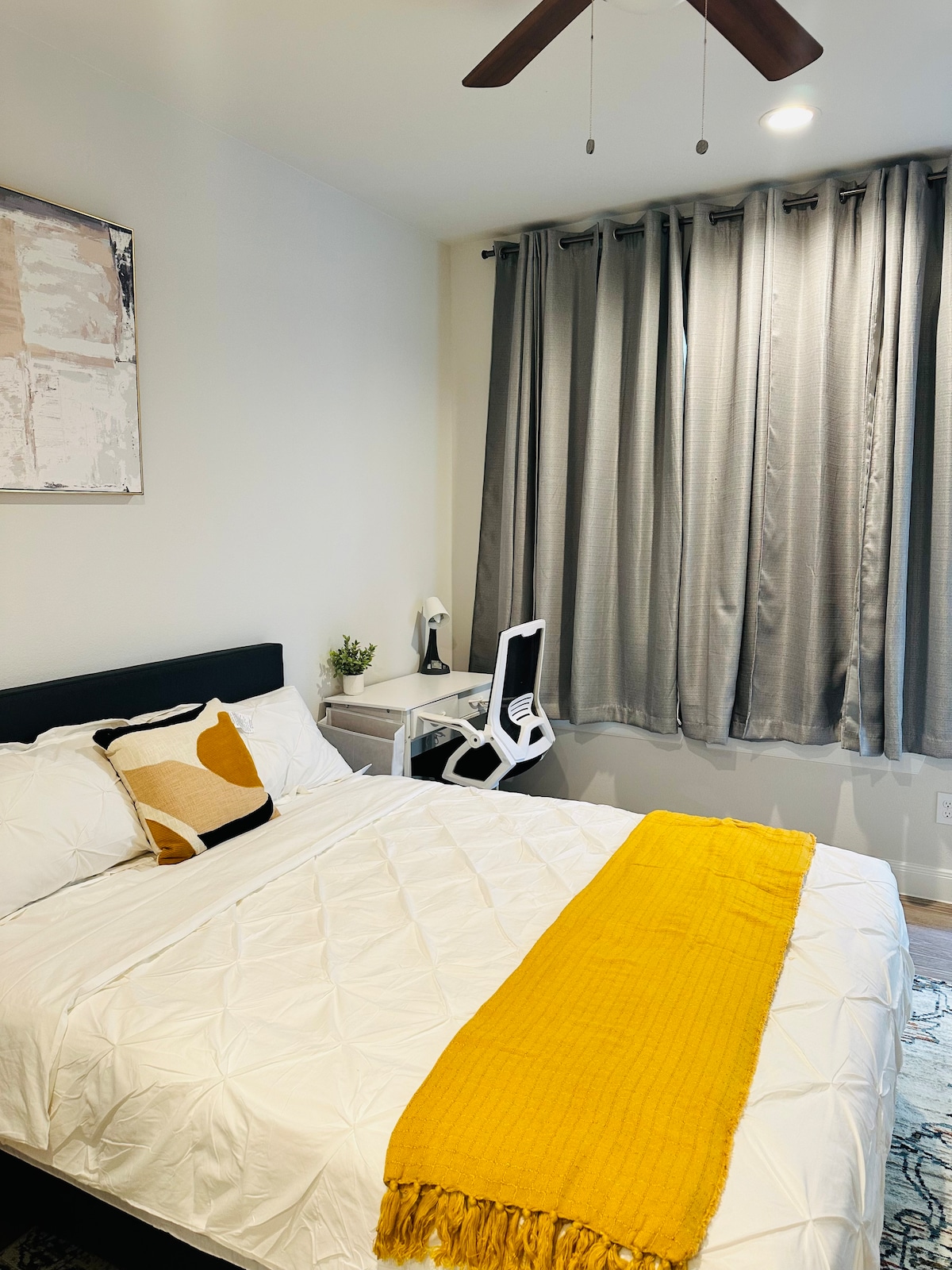 A serene bedroom is presented with a spacious bed featuring a white quilt and accent pillows. A vibrant yellow throw blanket is draped across the bed. Adjacent to the bed is a small desk with a modern chair, while floor-to-ceiling gray curtains frame the window.