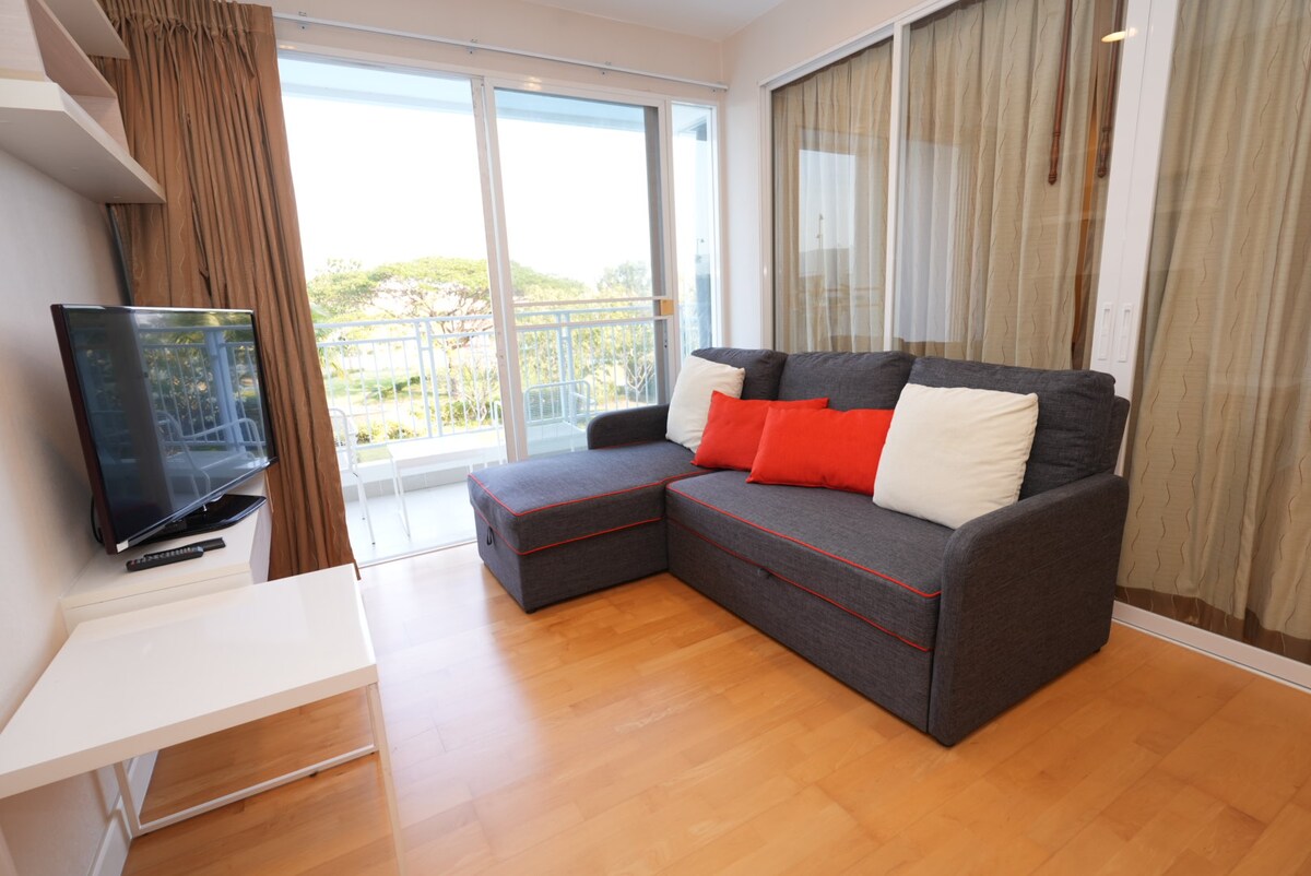 A comfortable living area is presented with a grey sectional sofa, accented by red and white cushions. A television is mounted on a sleek white stand. Floor-to-ceiling windows allow natural light to fill the space, with views of the surroundings partially obscured by translucent drapes.