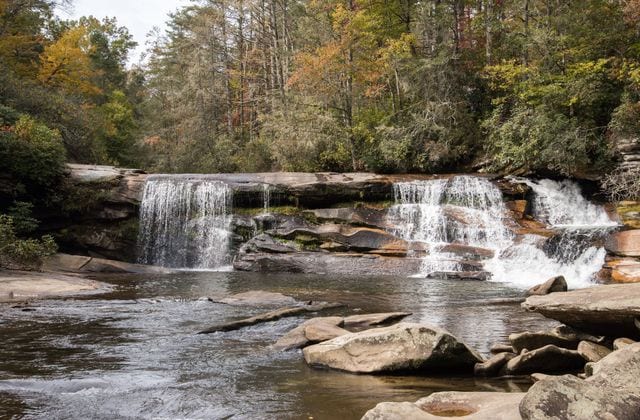A serene waterfall cascades over a rocky ledge into a gently flowing stream. Surrounding greenery features trees displaying autumn colors, creating a calming natural setting. Smooth stones are visible along the water's edge, inviting exploration of the tranquil environment.