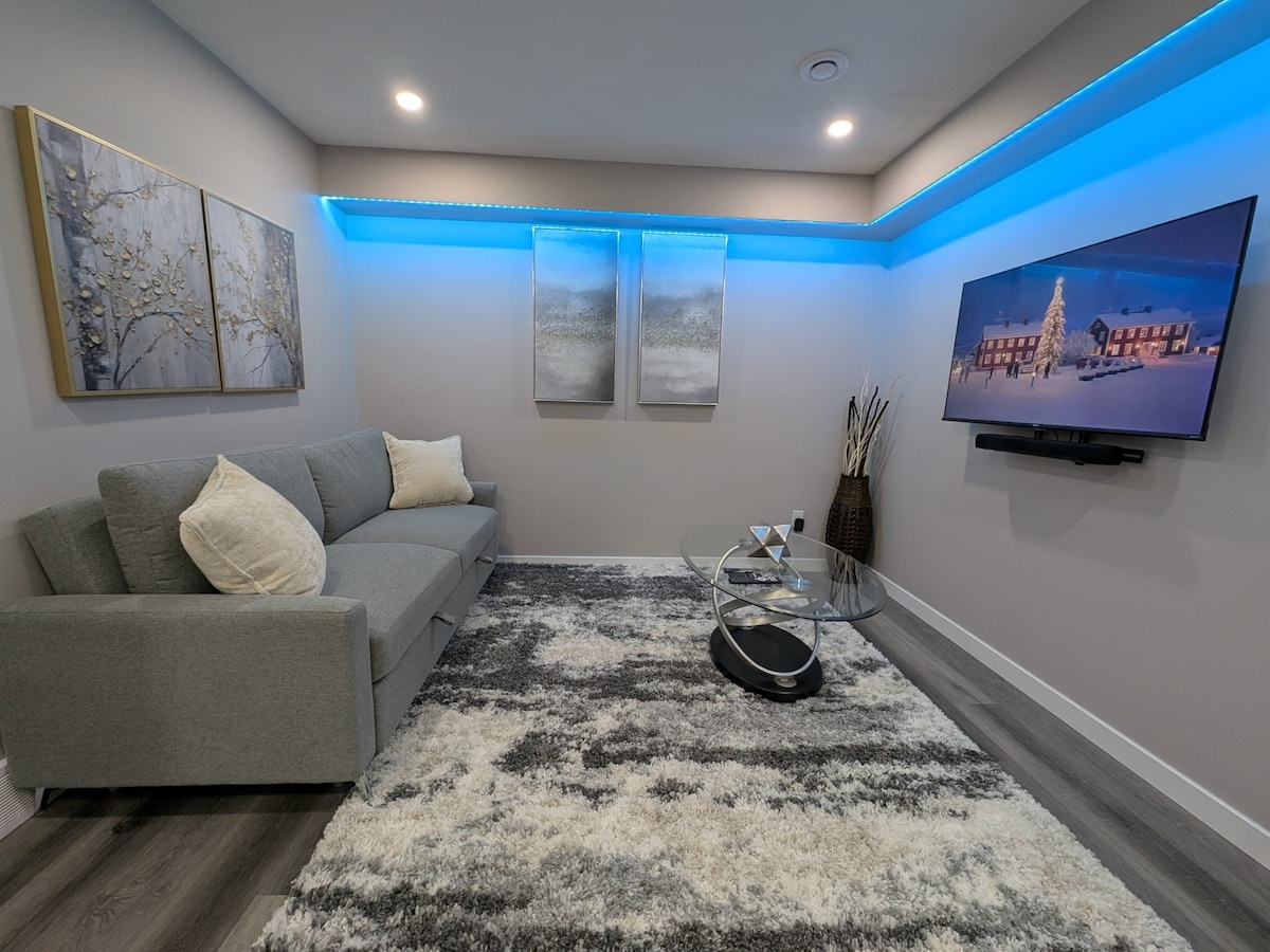 A modern living area features a cozy grey sofa adorned with plush throw pillows. Above, two framed artworks add a touch of elegance, while ethereal blue LED lighting enhances the ambiance. A glass coffee table is positioned on a soft area rug, paired with a wall-mounted TV displaying a scenic image.