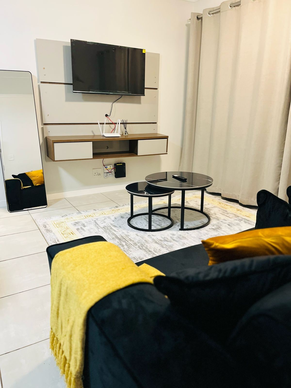 A cozy living area features a sleek black sofa adorned with decorative cushions and a soft yellow throw. Two circular black tables are positioned on a light-colored rug. A flat-screen TV is mounted above a minimalist shelf, and floor-length curtains frame the window.