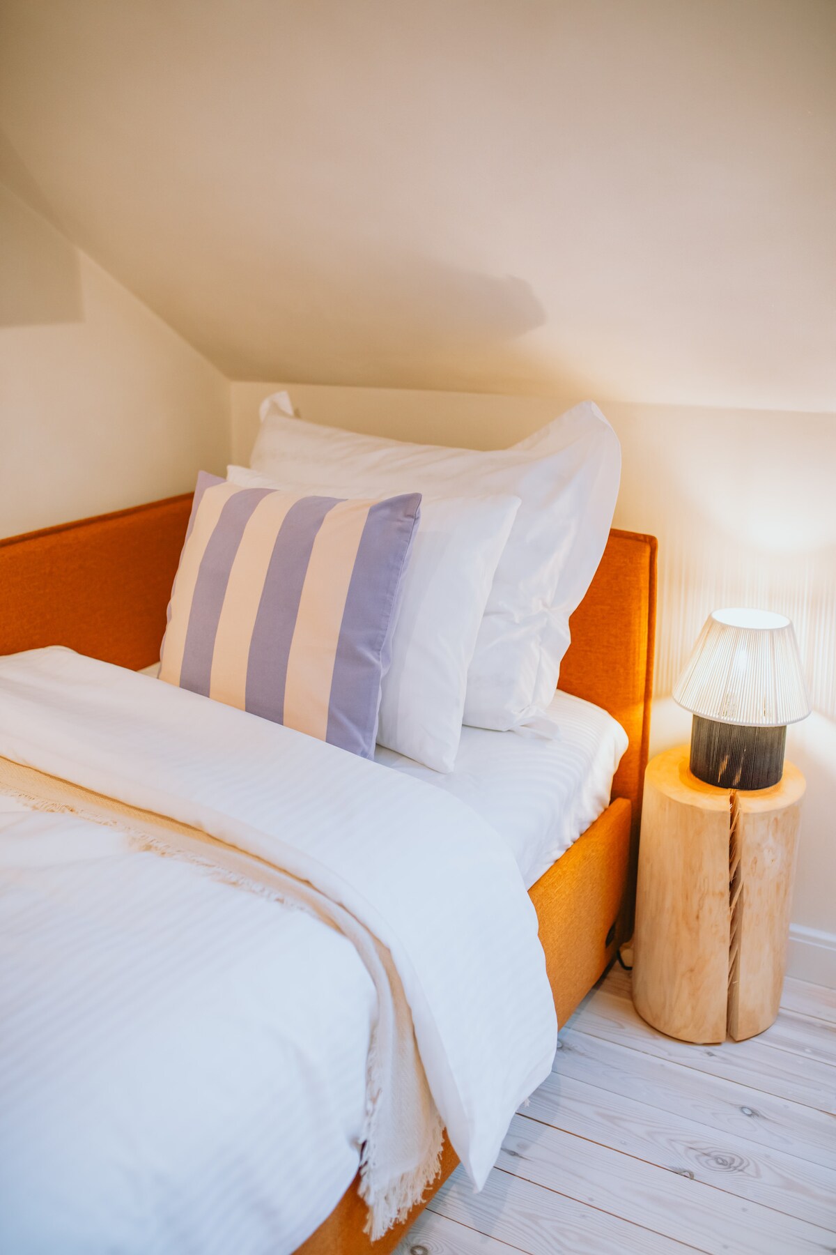 An inviting sleeping area features a spacious orange bed with striped and solid white pillows. A soft, white blanket is neatly folded. A wooden bedside table holds a stylish lamp that emits a gentle light, complementing the warm color palette of the room.