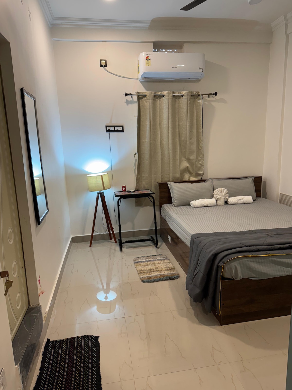 The bedroom features a spacious bed with neatly arranged bedding and a decorative element atop. A bedside table holds a lamp, and a full-length mirror is mounted on the wall. A curtain frames a window, while a small rug adds texture to the floor.