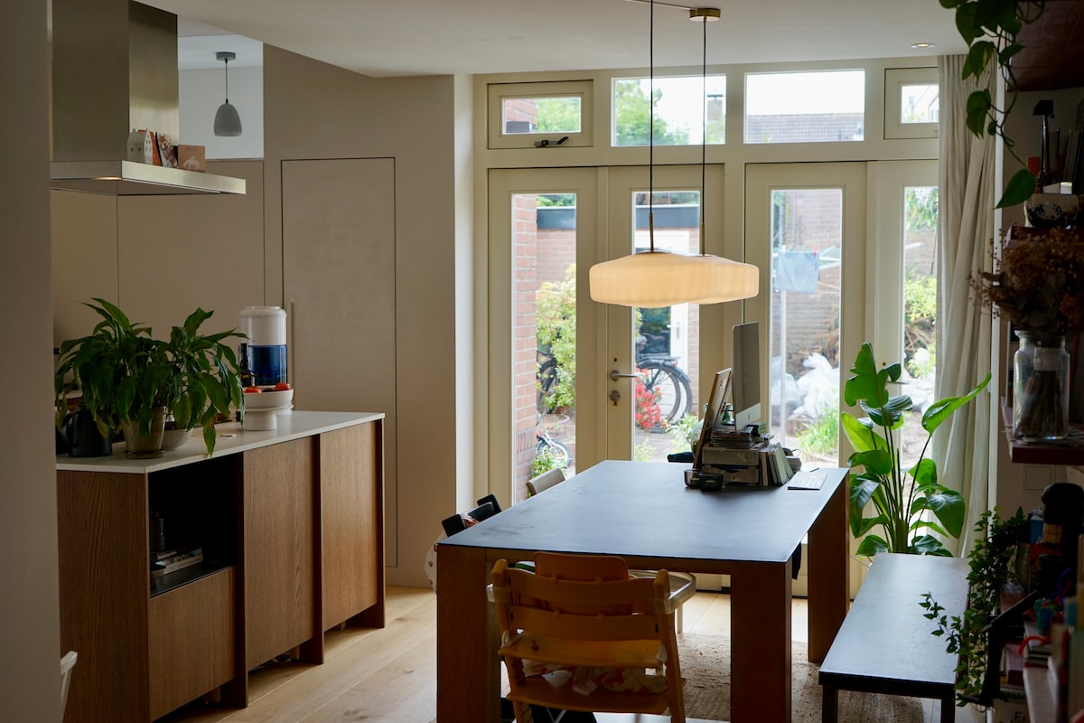 A modern kitchen area features a light wooden table accompanied by a few chairs. Large glass doors invite natural light and provide a view of the garden. Potted plants add greenery, and a stylish pendant light hangs above the table, enhancing the functional aesthetic of the space.