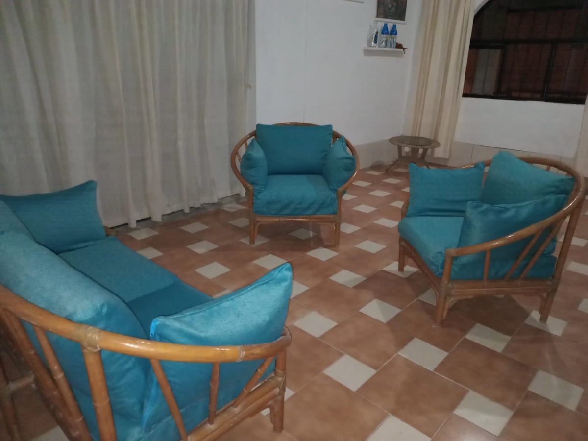 A seating area is presented with two armchairs and a matching two-seater sofa, all upholstered in turquoise fabric. The floor features tiled patterns in warm tones, and sheer curtains allow for gentle light to filter through from the window.