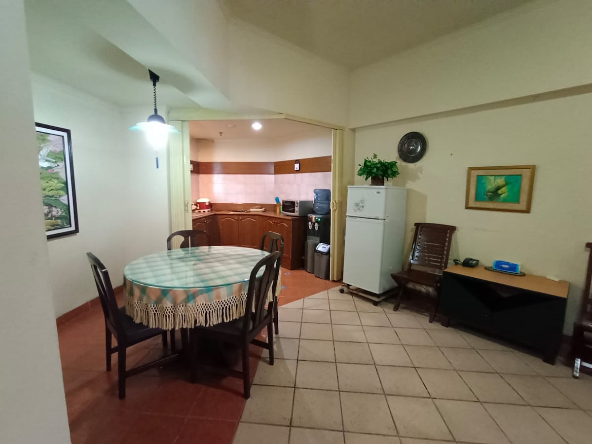 A dining area is featured with a round table surrounded by six wooden chairs. A small kitchen is visible in the background, equipped with a refrigerator and various appliances. Soft lighting is provided by an overhead pendant lamp in a space that offers a warm, inviting atmosphere.