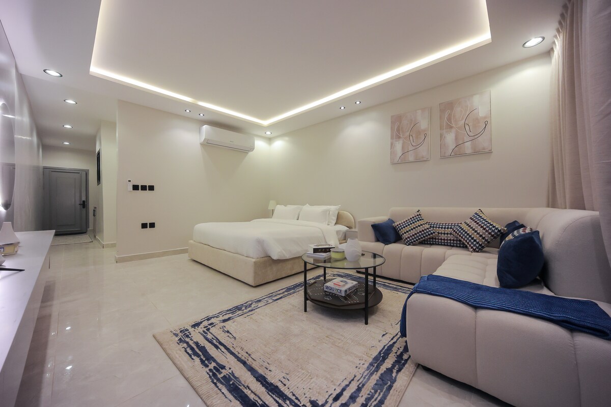 A spacious room features a large bed with soft bedding and a contemporary sofa, positioned next to a round coffee table. Natural light filters in through elegantly draped curtains. Wall art complements the minimalist decor, while recessed lighting adds a modern touch.