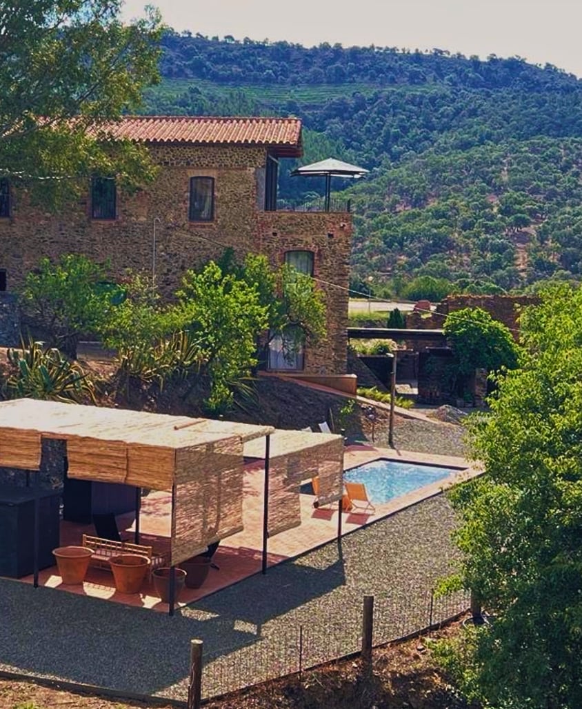 A rustic stone building is complemented by a sunny terrace featuring lounge seating and an inviting pool. Surrounding greenery and hills enhance the natural setting, while a large umbrella provides shade over the outdoor area, creating a serene atmosphere.