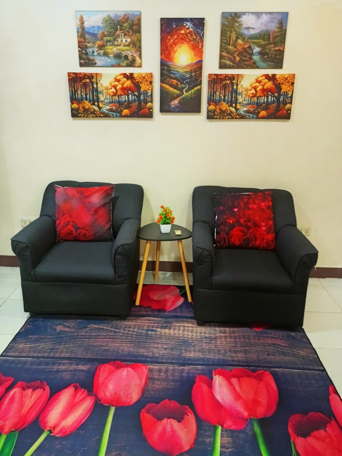 Two black armchairs are positioned opposite each other, accompanied by a small round table. Colorful wall art featuring landscapes and floral designs decorates the walls above. A rug displaying vibrant red tulips anchors the space, contributing to a cozy and welcoming atmosphere.