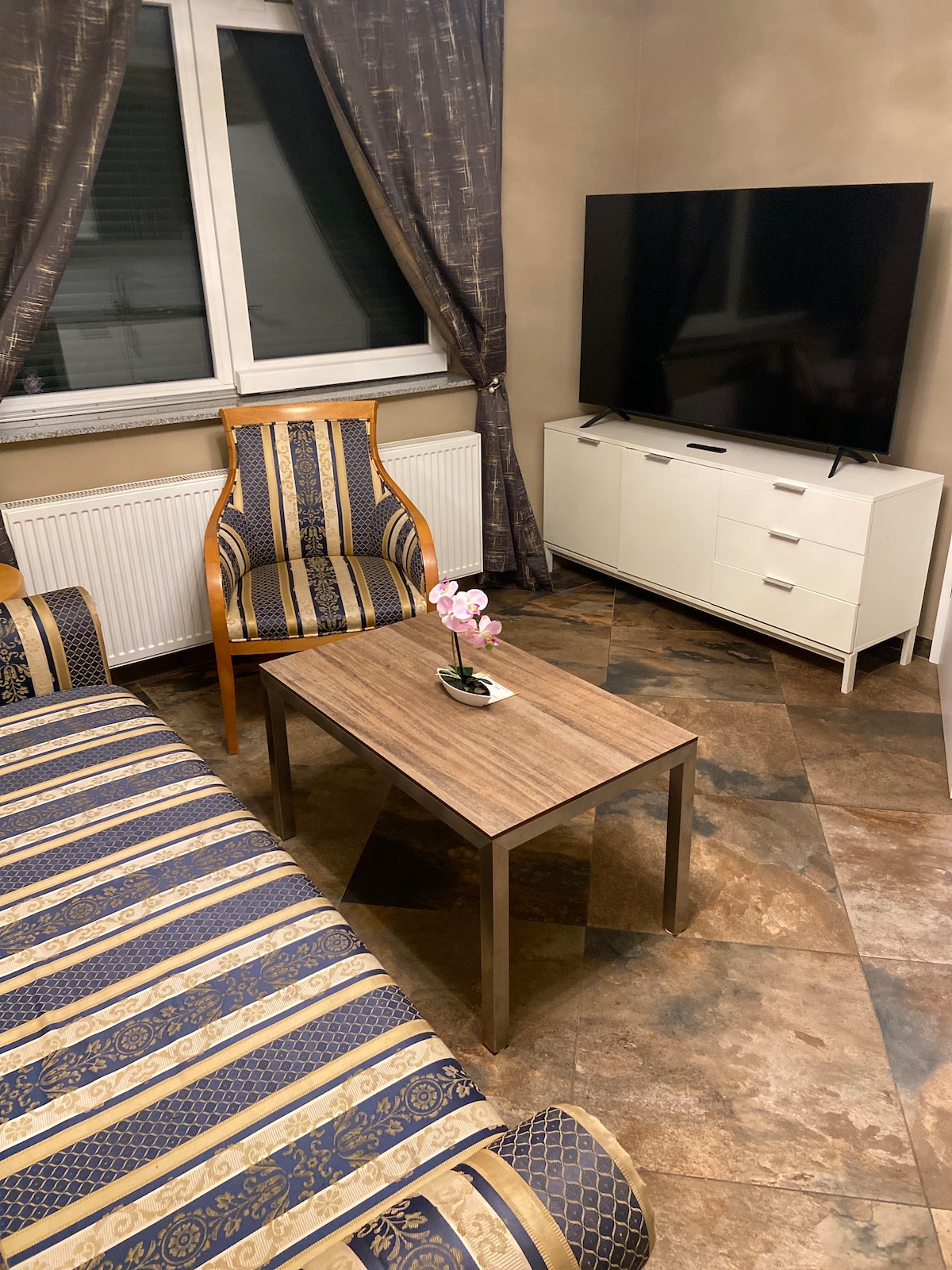 A cozy seating area is presented with two upholstered chairs featuring a subtle striped pattern. A wooden coffee table sits centrally, adorned with a small potted plant. A sleek television is positioned against a light-colored wall, while drapes frame the window.
